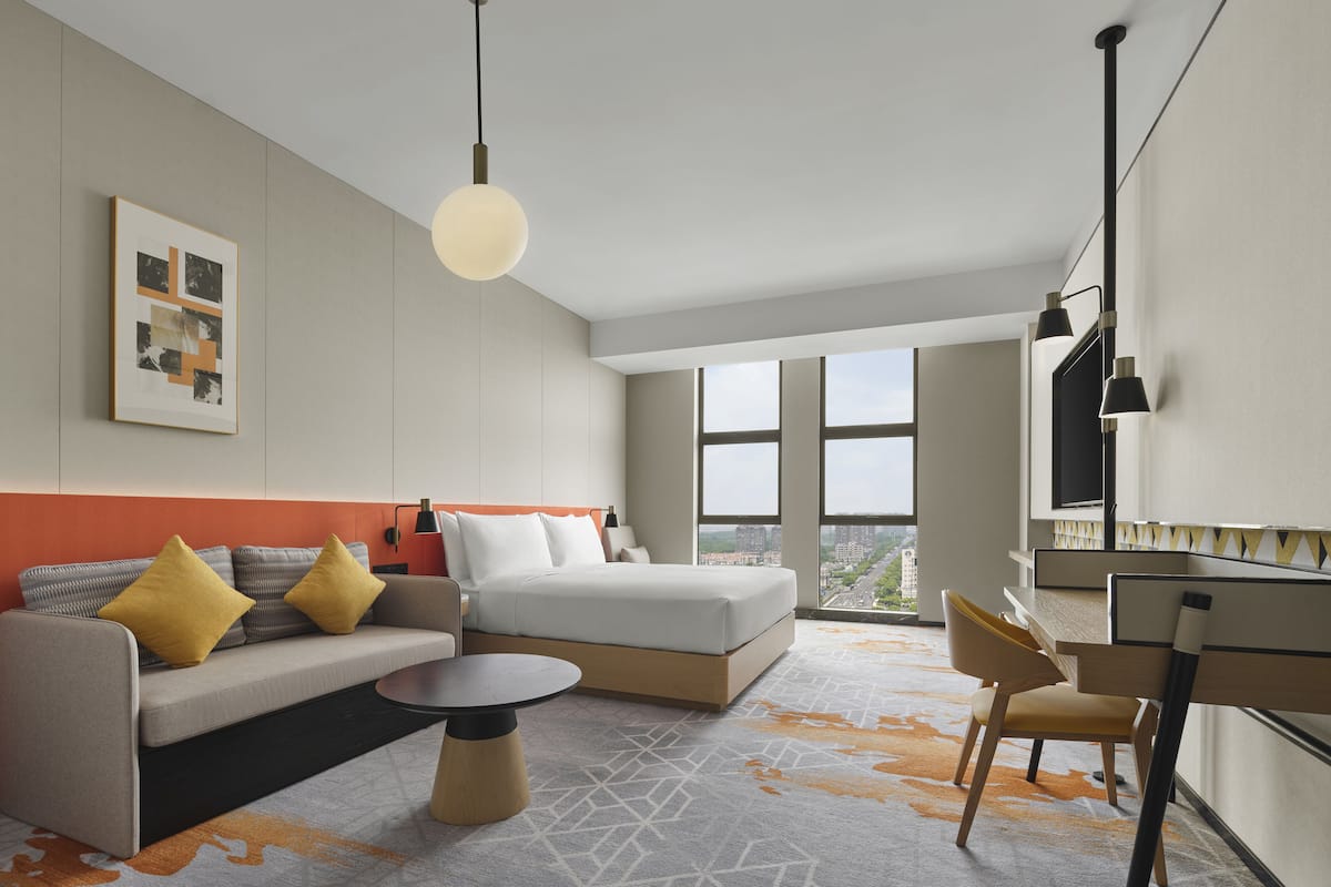 Ein modernes Hotelzimmer mit Sofa, rundem Couchtisch, Schreibtisch und großem Fenster mit Blick auf die Stadt.