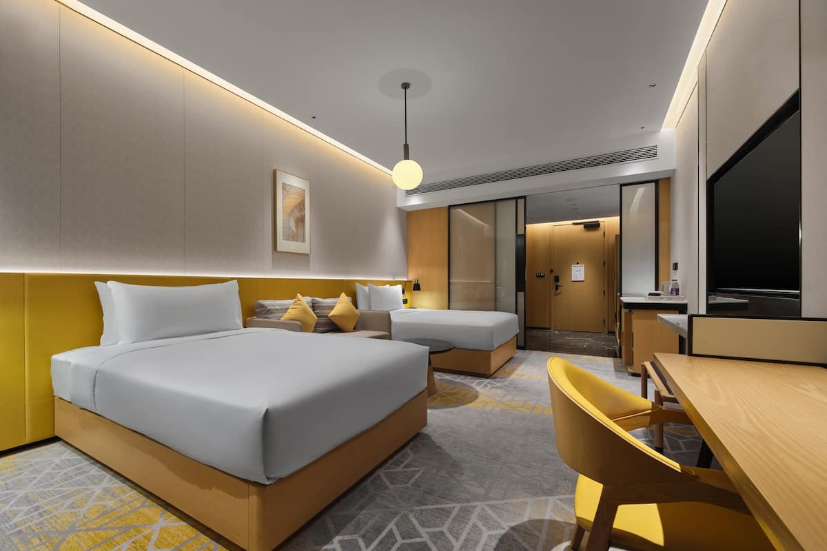 Ein modernes Hotelzimmer mit einem großen Bett, einem Schreibtisch und einem Sessel. Es gibt einen Fernseher, eine Schreibtischlampe und ein gerahmtes Bild an der Wand.