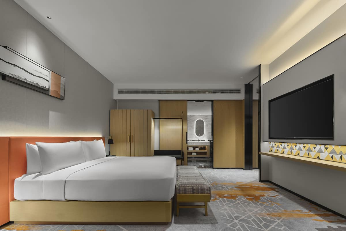 Ein modernes Hotelzimmer mit einem großen Bett, einem Fernseher, einem Kleiderschrank und einem durch eine offene Tür einsehbaren Badezimmer.
