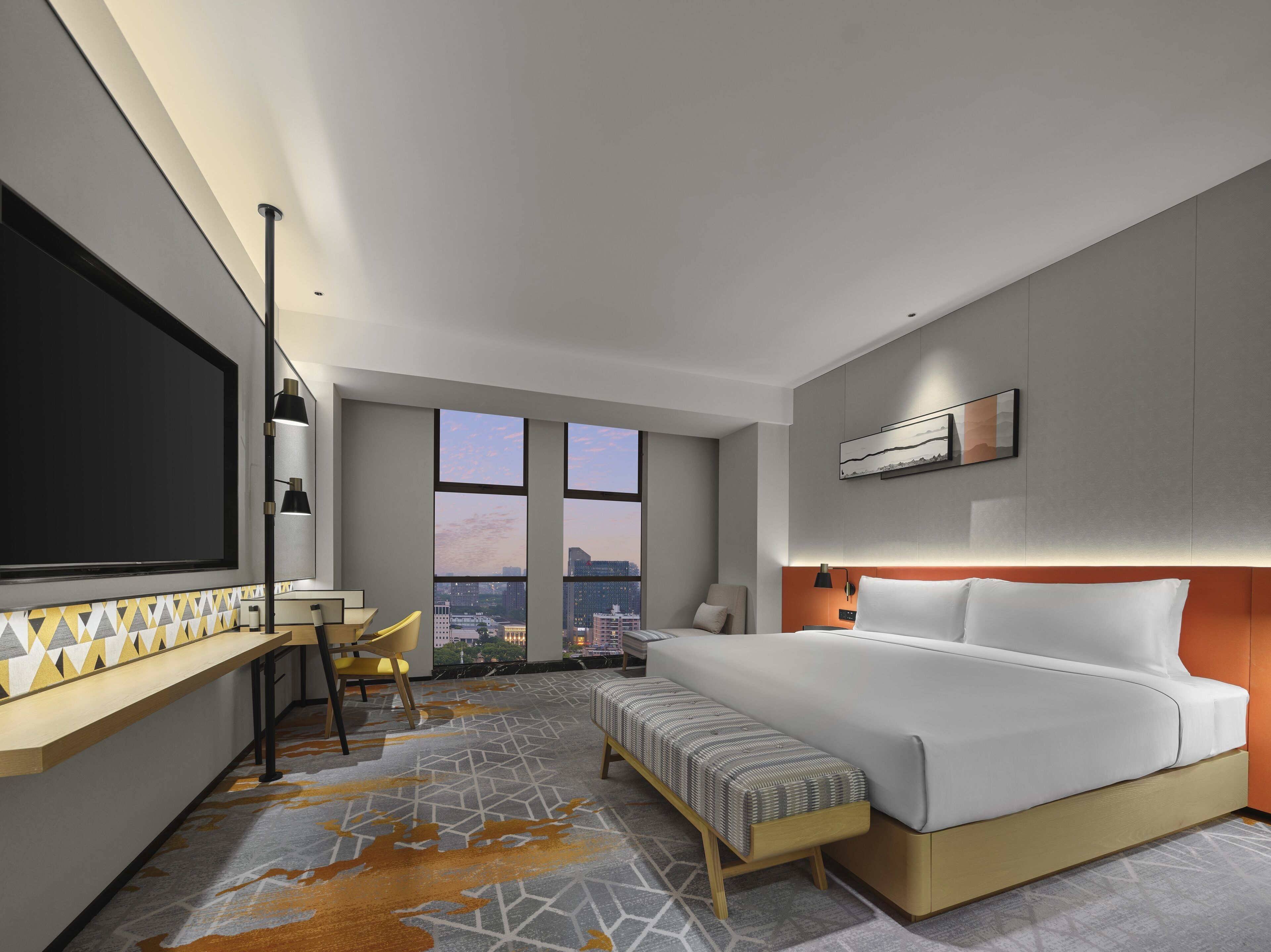 A modern hotel room with a large bed, a desk with a chair, a bench, and a large window offering a city view.