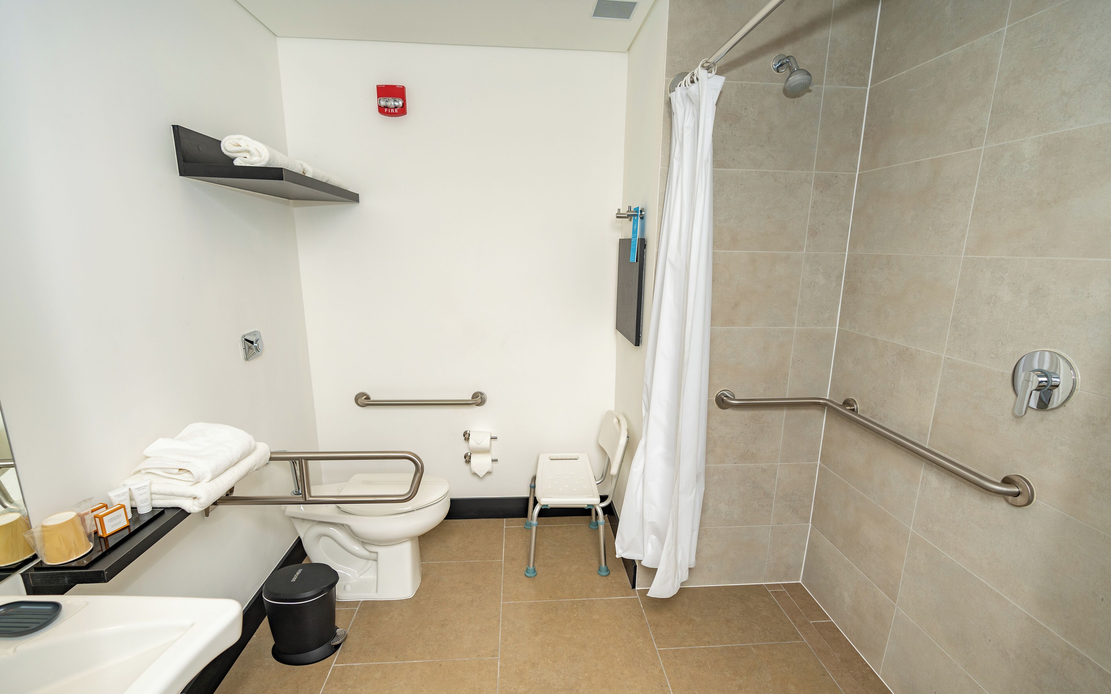 A bathroom with a toilet, handrails, a shower, and a waste bin.