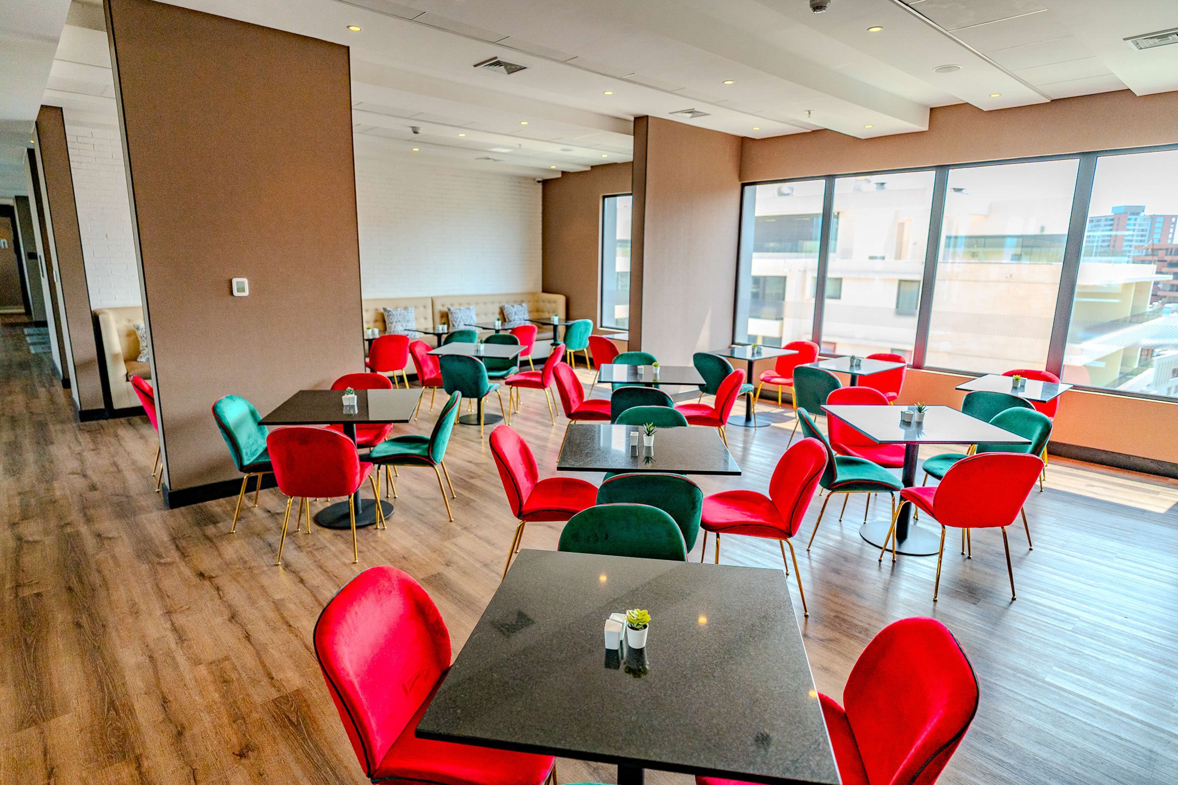 Un comedor moderno con sillas rojas y verdes, varias mesas y grandes ventanales con vistas a la ciudad.