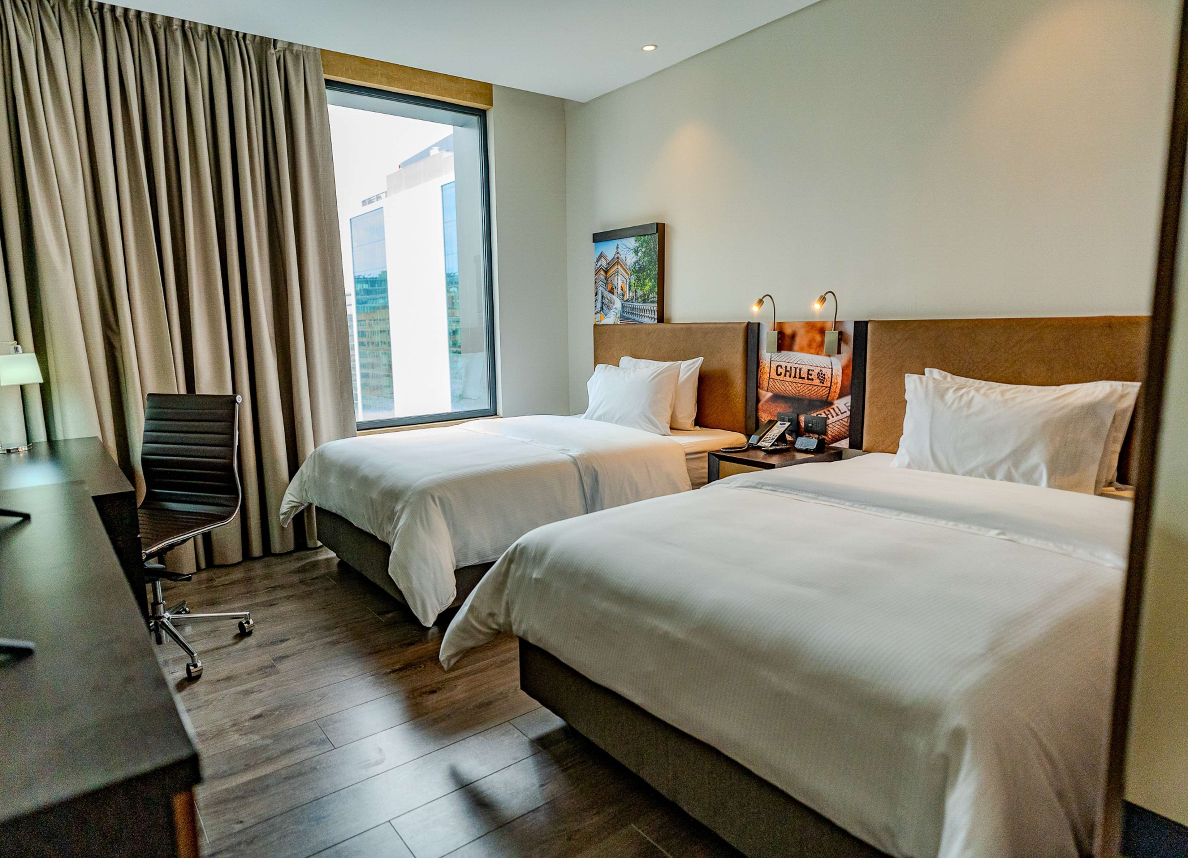 Een hotelkamer met twee bedden, een bureau, een stoel en een groot raam met gordijnen.