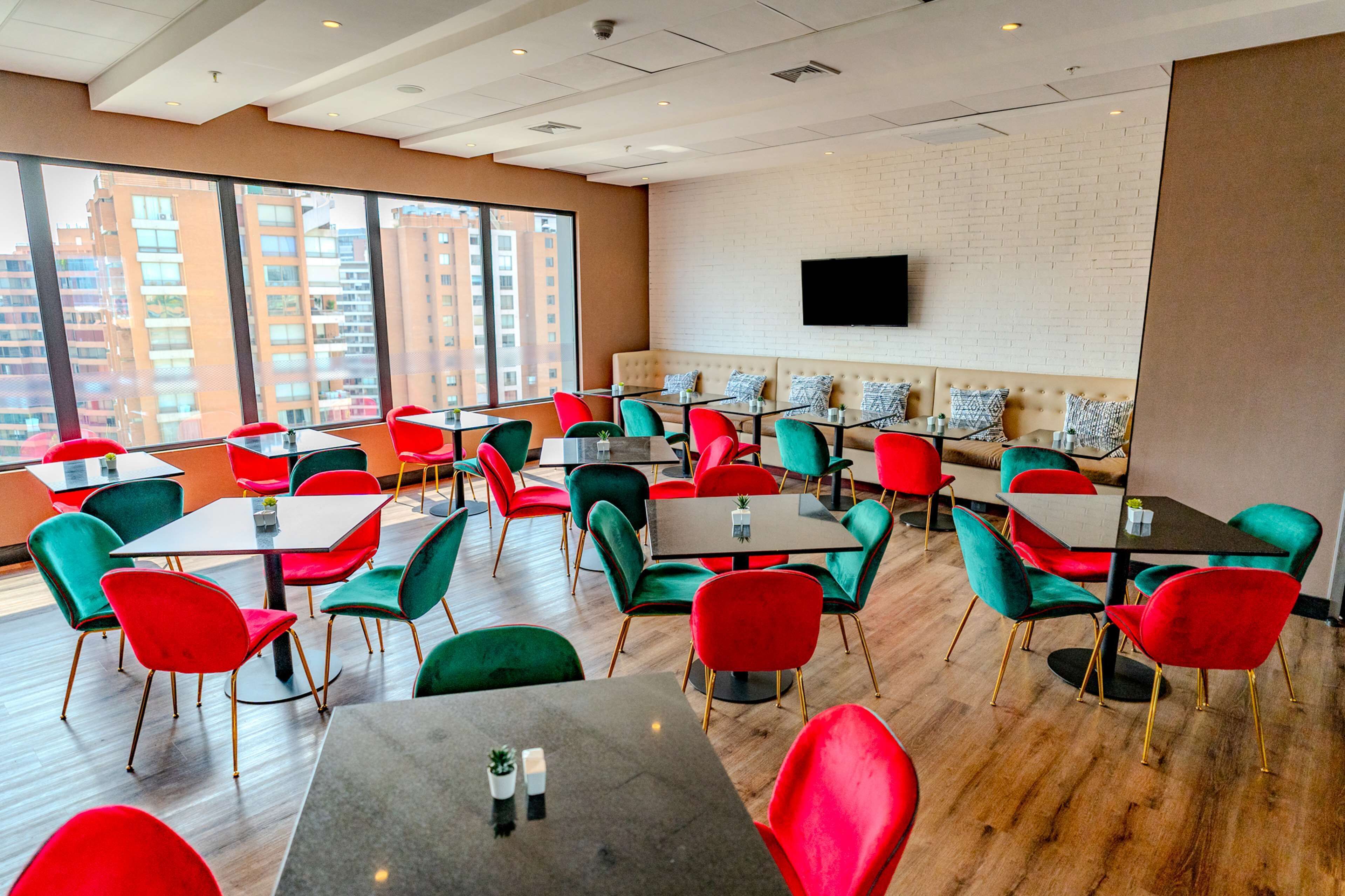 A modern dining area with red and green chairs, multiple tables, and a large window offering a city view.