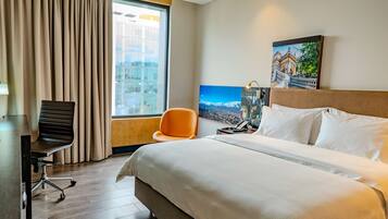 Quarto de hotel com uma cama grande, uma escrivaninha, uma cadeira laranja, uma janela com cortinas e uma pintura de paisagem urbana na parede.