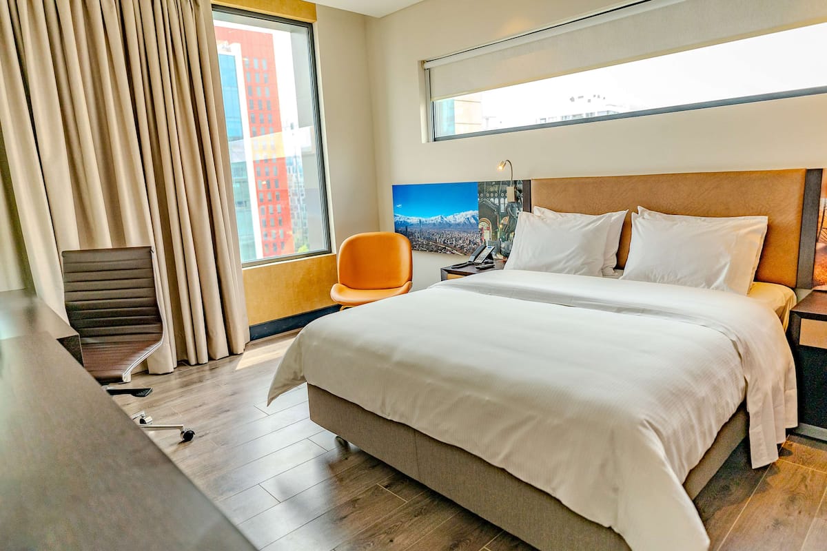 Une chambre d’hôtel avec un grand lit, un bureau, un fauteuil orange, une fenêtre donnant sur le paysage urbain et un tableau accroché au mur.