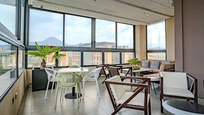 Een ruim balkon met een tafel en stoelen, grote ramen met uitzicht op de stad, en potplanten.