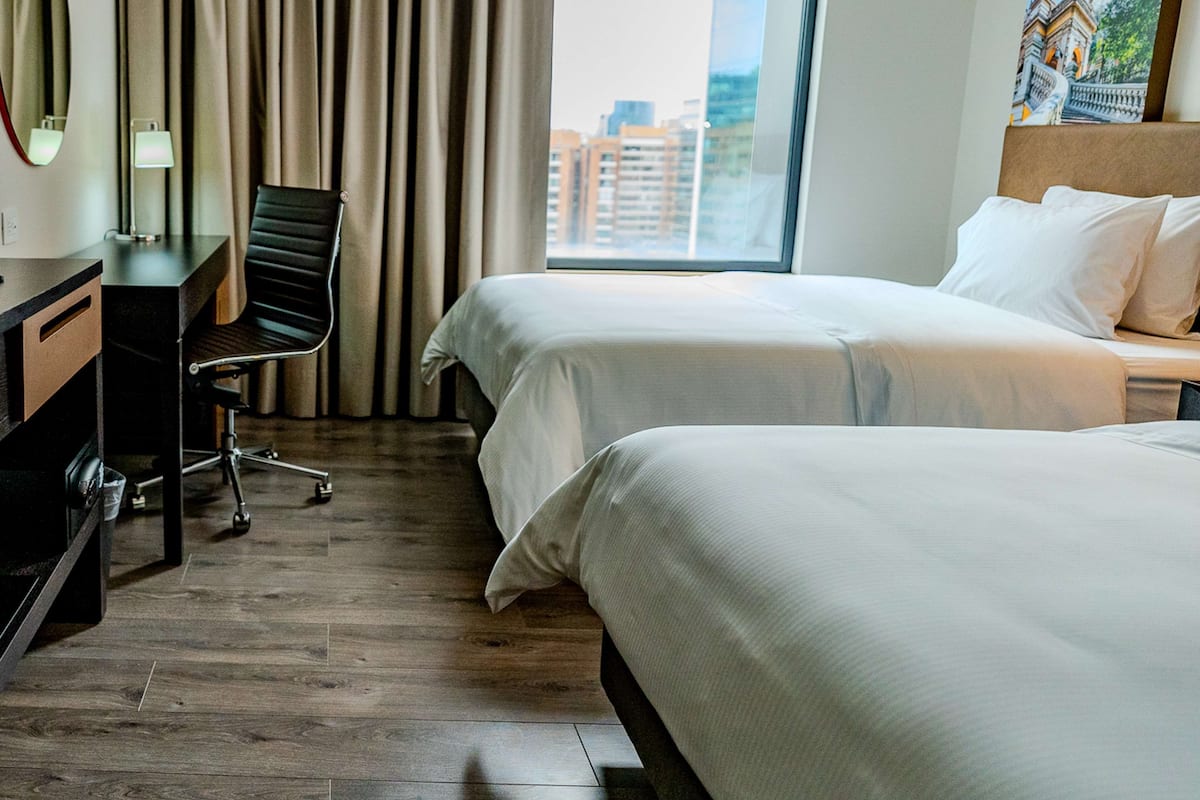 Une chambre d’hôtel avec deux lits, un bureau, une chaise, une fenêtre donnant sur la ville et un tableau au mur.