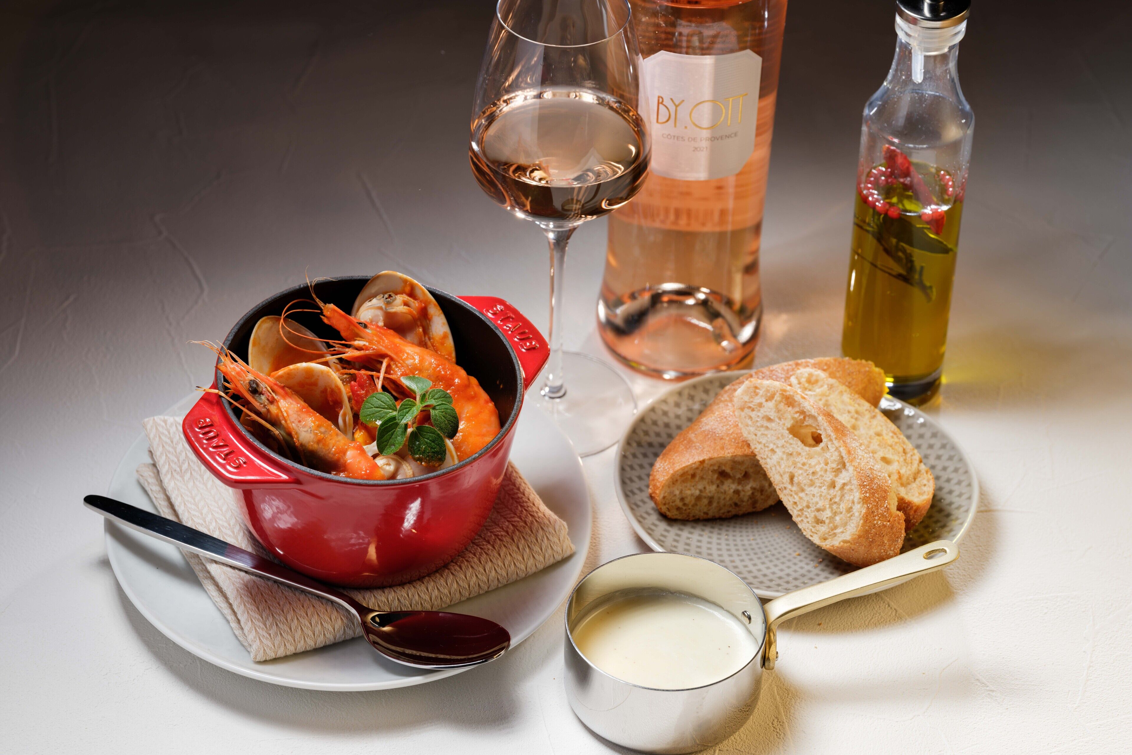 Une table à manger avec un plat rouge contenant des fruits de mer, un verre de vin, une bouteille d’huile d’olive, un petit bol de sauce, une cuillère, un couteau et une tranche de pain sur une assiette.