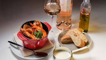 Une table à manger avec un plat rouge contenant des fruits de mer, un verre de vin, une bouteille d’huile d’olive, un petit bol de sauce, une cuillère, un couteau et une tranche de pain sur une assiette.