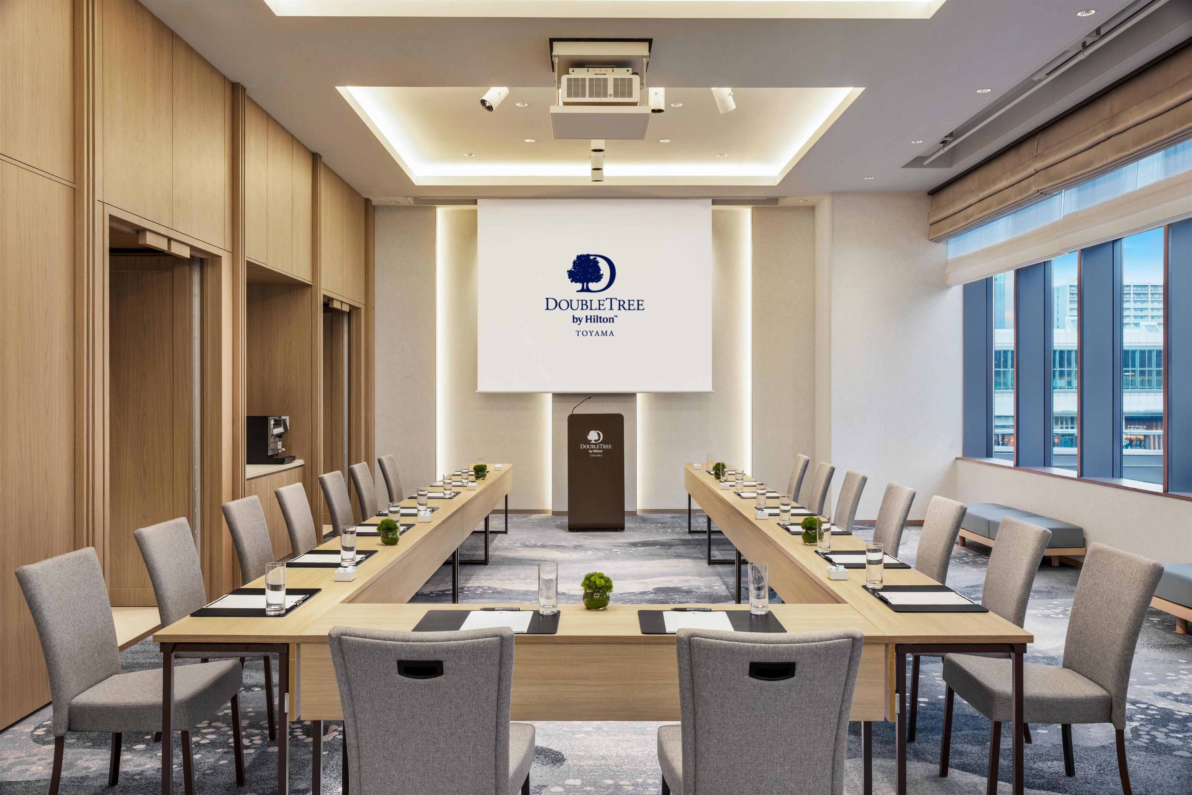 A conference room with a long table, chairs, a projector screen, and a logo on the wall.