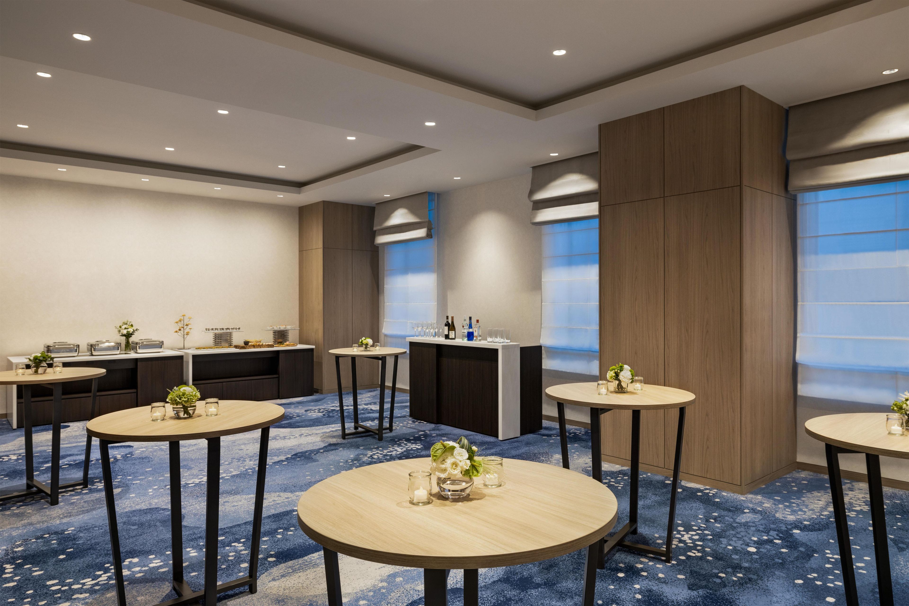 A hotel room with a dining area featuring round tables, a buffet setup, and a large window with blinds.
