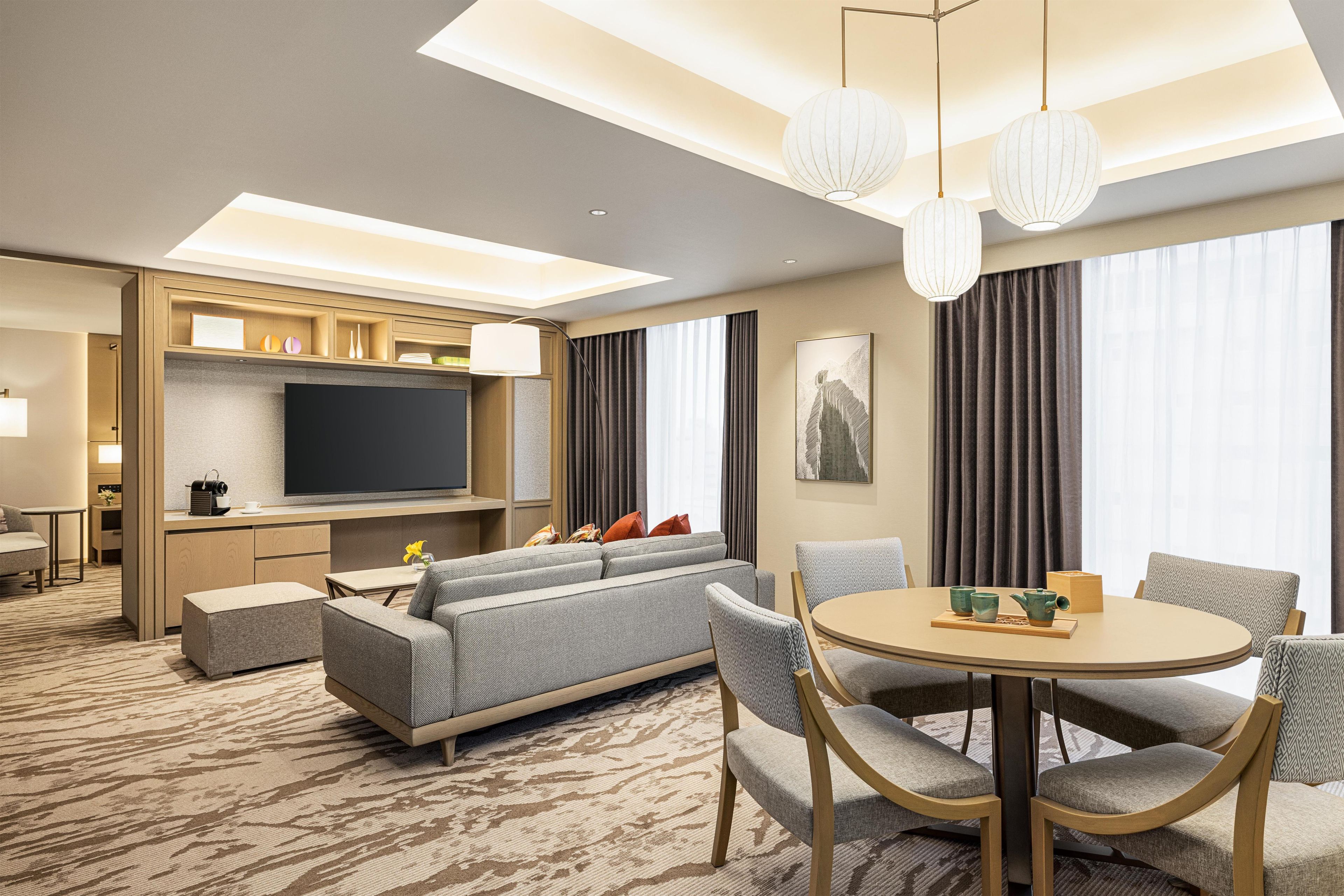 A modern hotel room with a flat-screen TV, a sofa, a dining table with chairs, and a large window with curtains.