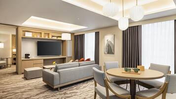 A modern hotel room with a flat-screen TV, a sofa, a dining table with chairs, and a large window with curtains.