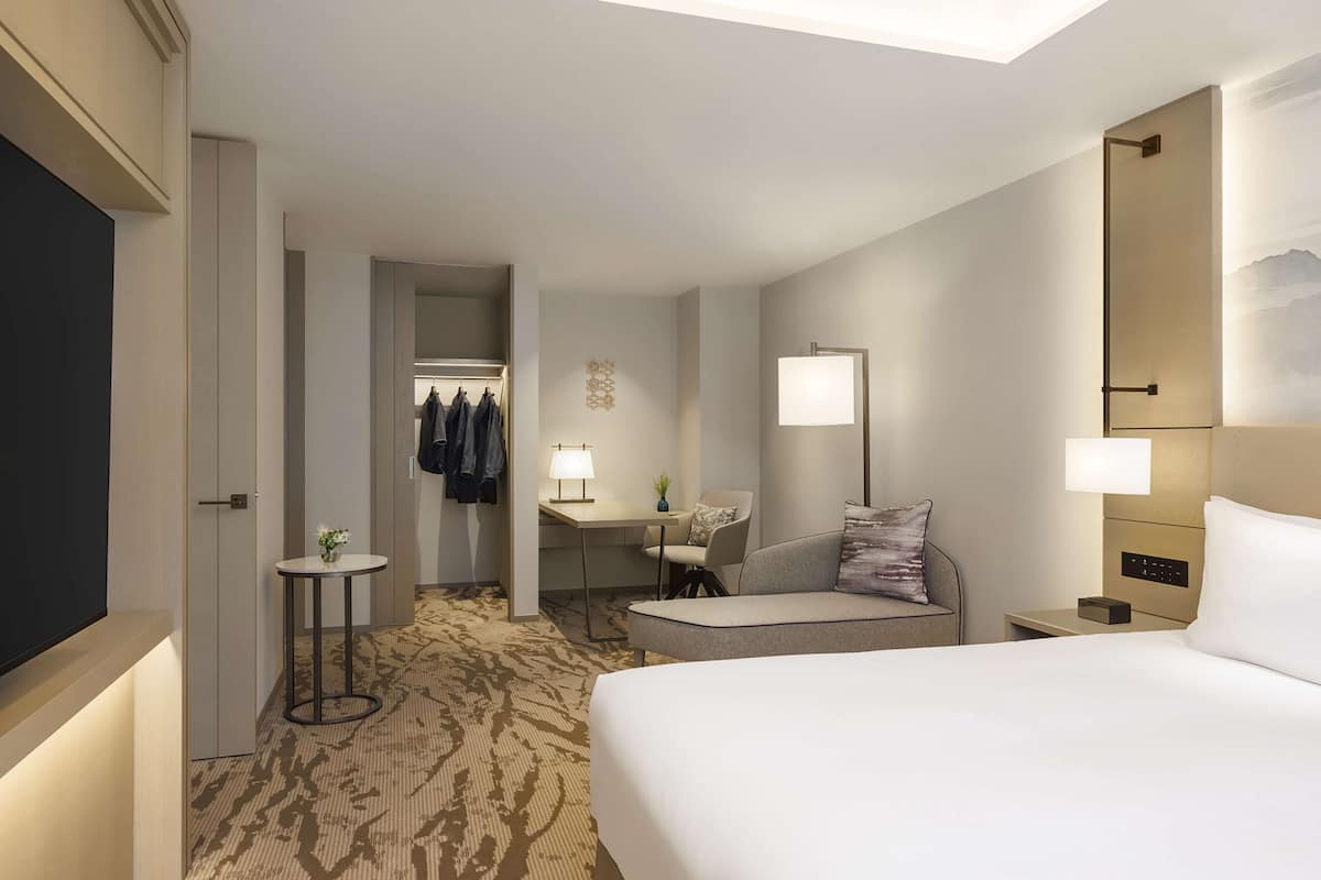 A hotel room with a large bed, a desk with a lamp, a chair, a small table with a plant, and a coat rack with jackets.