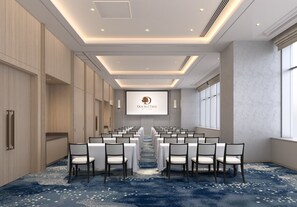 Une salle de conférence avec des rangées de tables et de chaises blanches, un écran de projection portant le logo « DOUBLETREE » et une moquette bleue à motifs blancs.