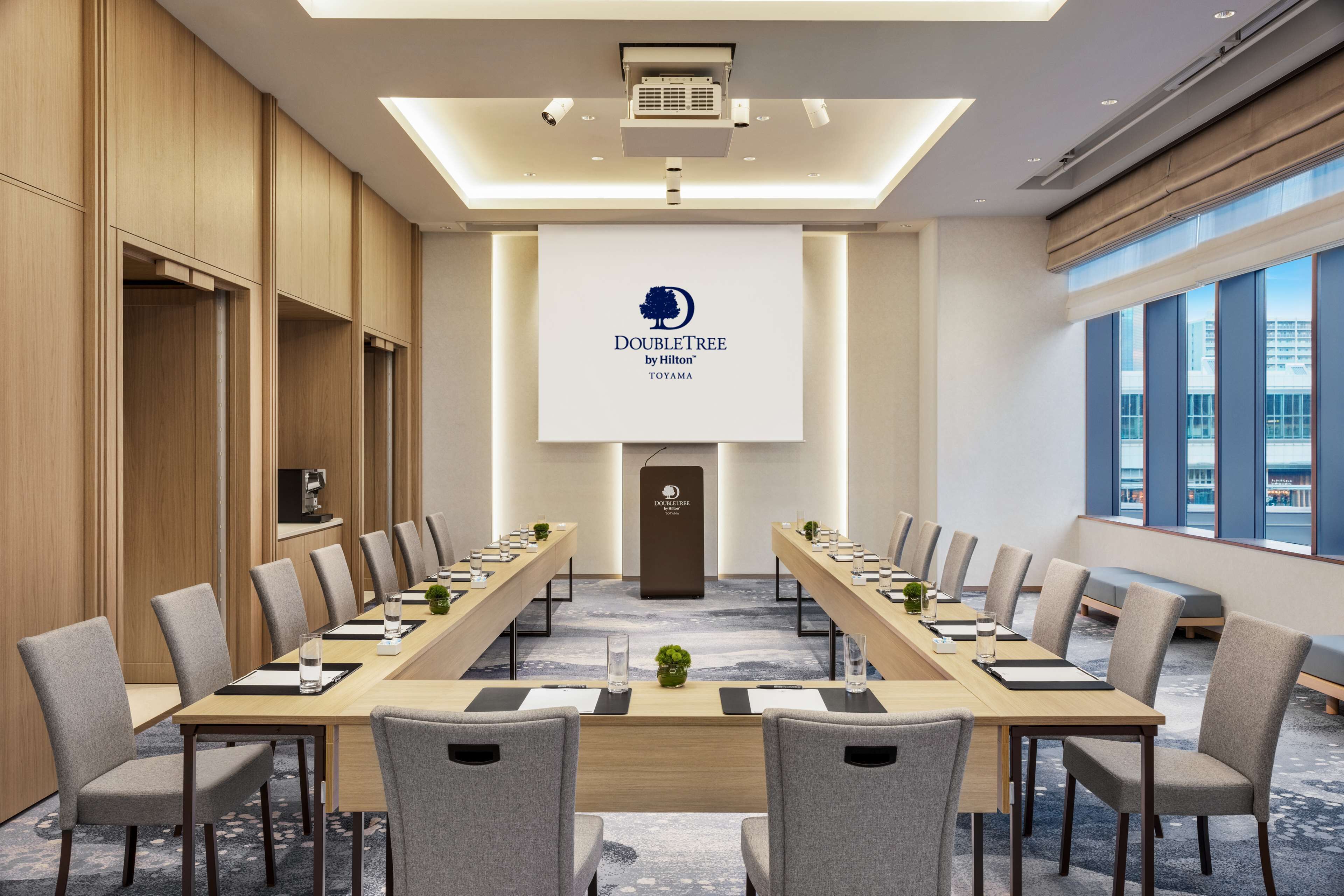 A conference room with a long table, chairs, a podium, and a large screen displaying the DoubleTree by Hilton logo.