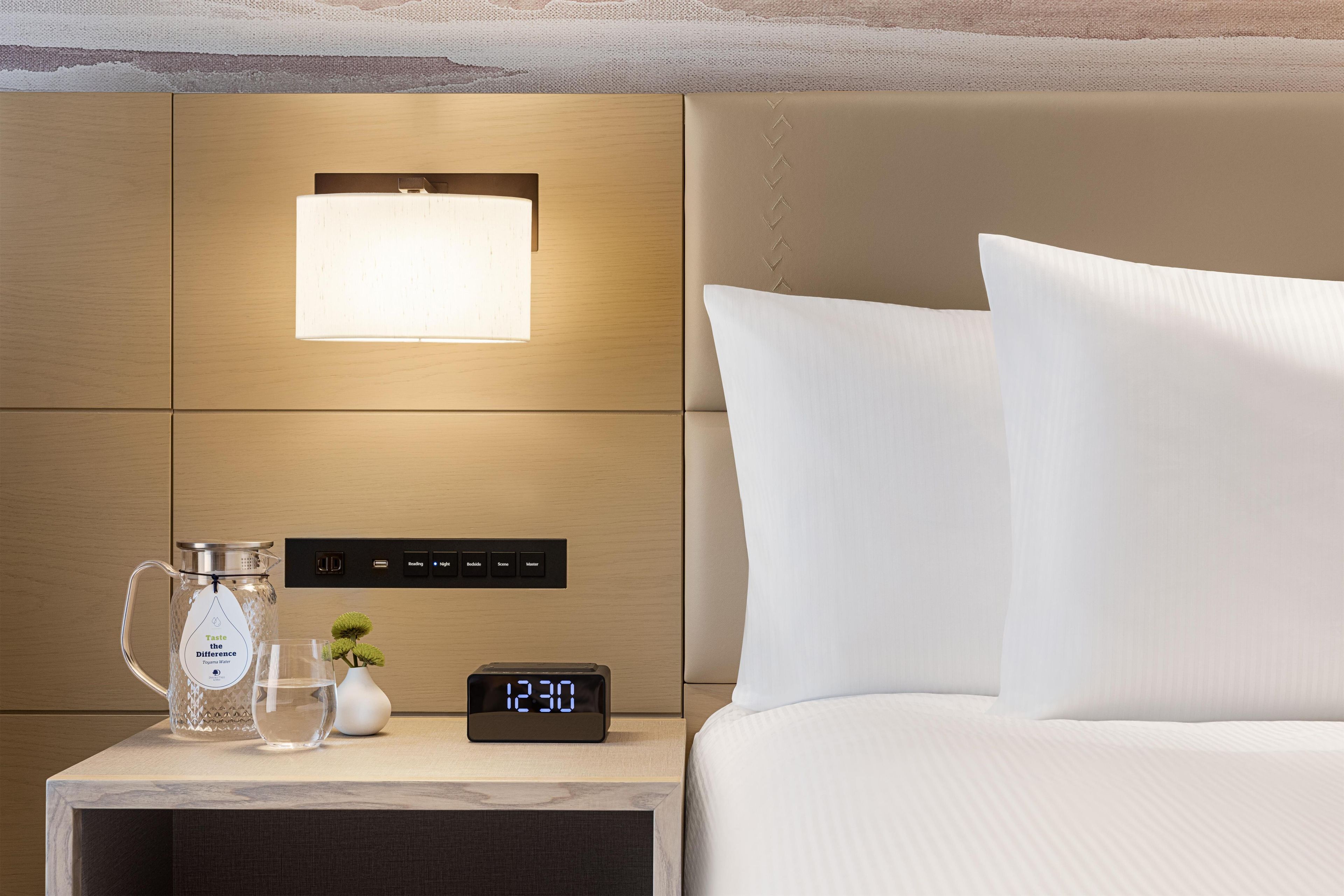 A neatly made hotel room with a bed, a bedside table, a nightstand, a digital clock, and a wall-mounted light fixture.