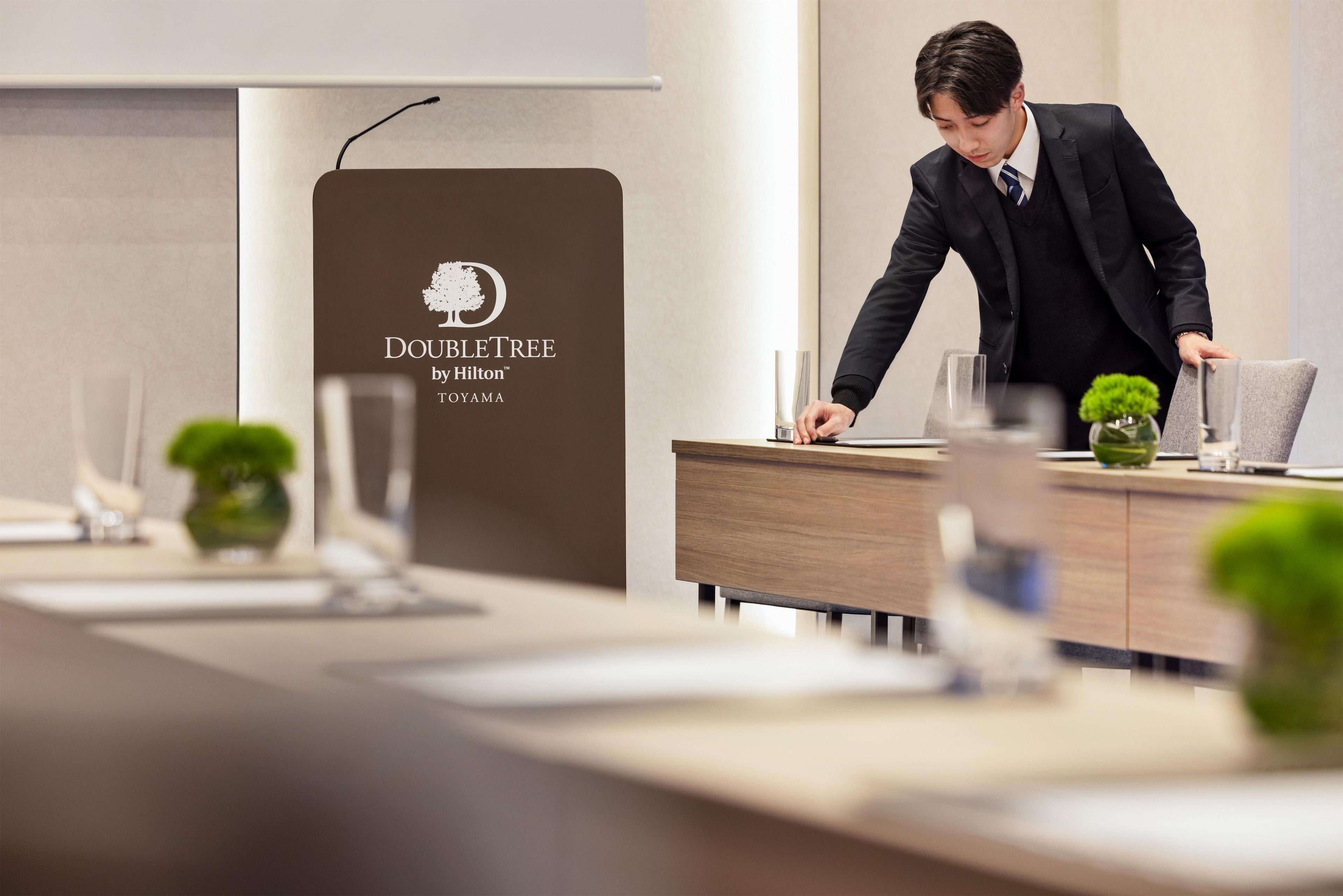 A hotel conference room with a podium displaying the DoubleTree by Hilton logo, a modern wooden desk, and a glass of water.