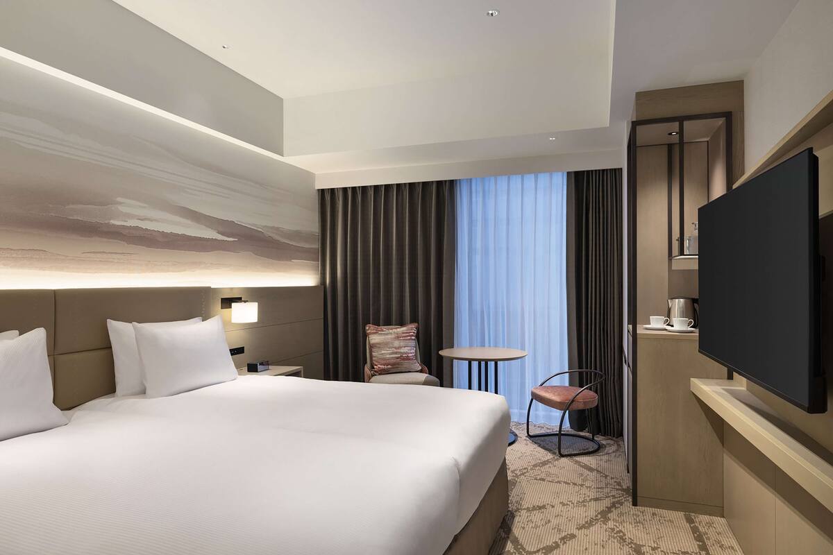 A modern hotel room with a large bed, a desk with a flat-screen TV, a small round table, and a chair.