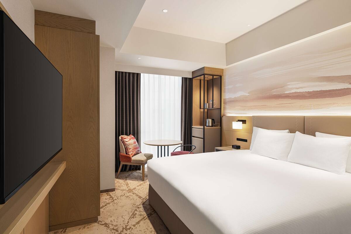 A hotel room with a large bed, a flat-screen TV, a seating area with a chair, a small table, and a bookshelf.