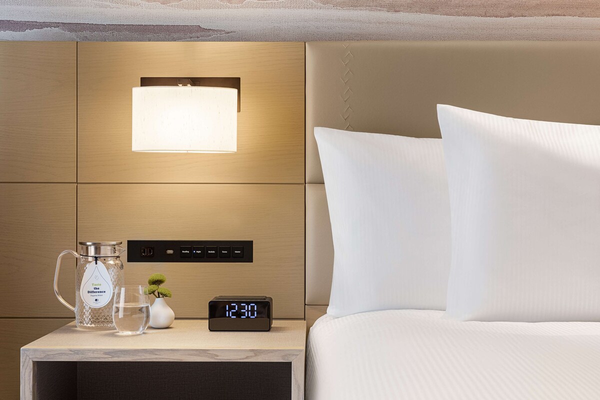 A neatly made hotel room with a bed, a bedside table with a digital clock, a glass of water, a vase with a plant, and a wall-mounted light.