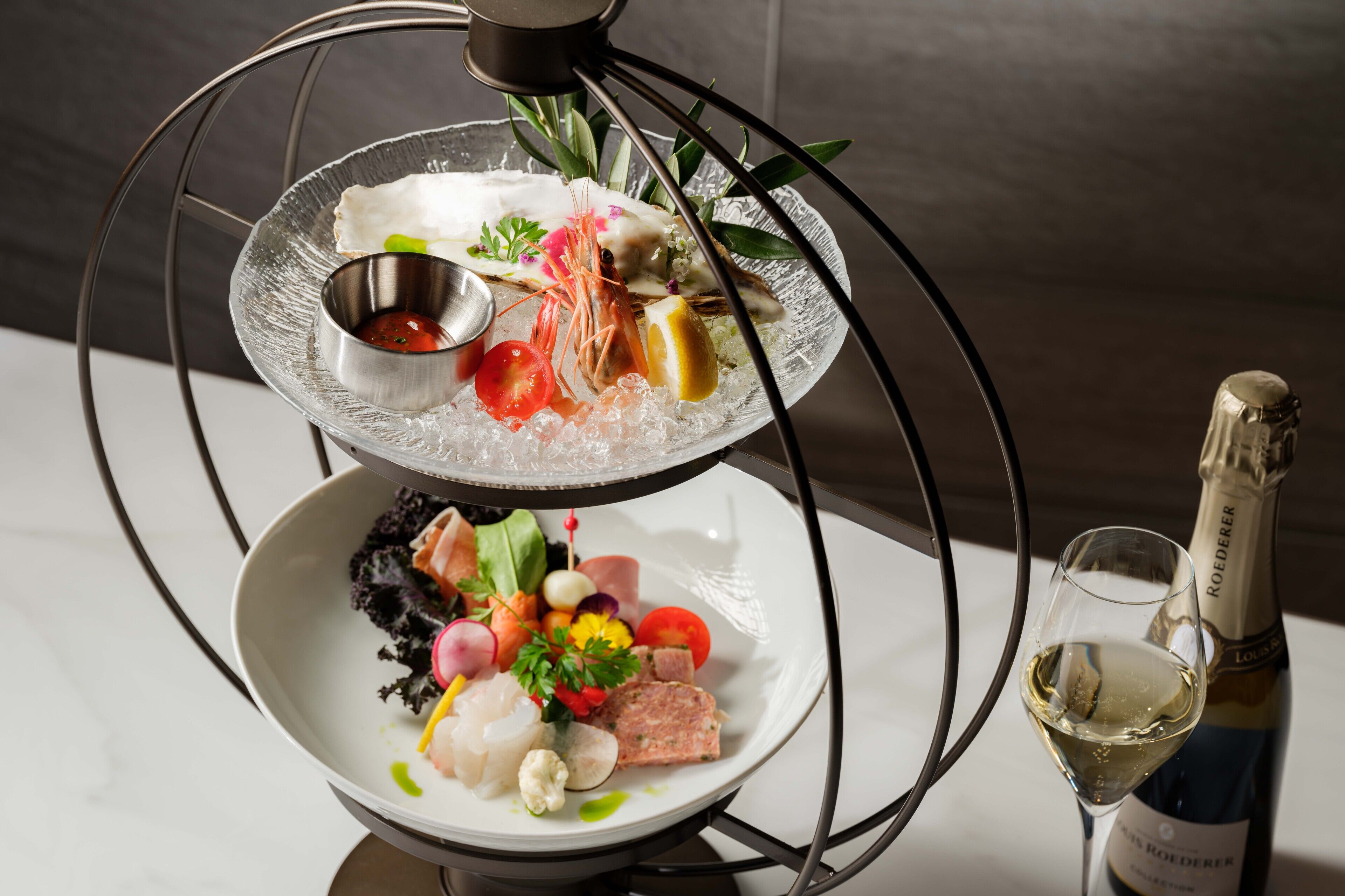 A multi-tiered glass dish with seafood and vegetables, a small bowl of sauce, and a bottle of wine.