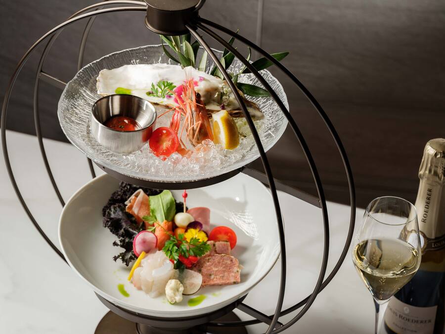 A multi-tiered glass dish with seafood and vegetables, a small bowl of sauce, and a bottle of wine.
