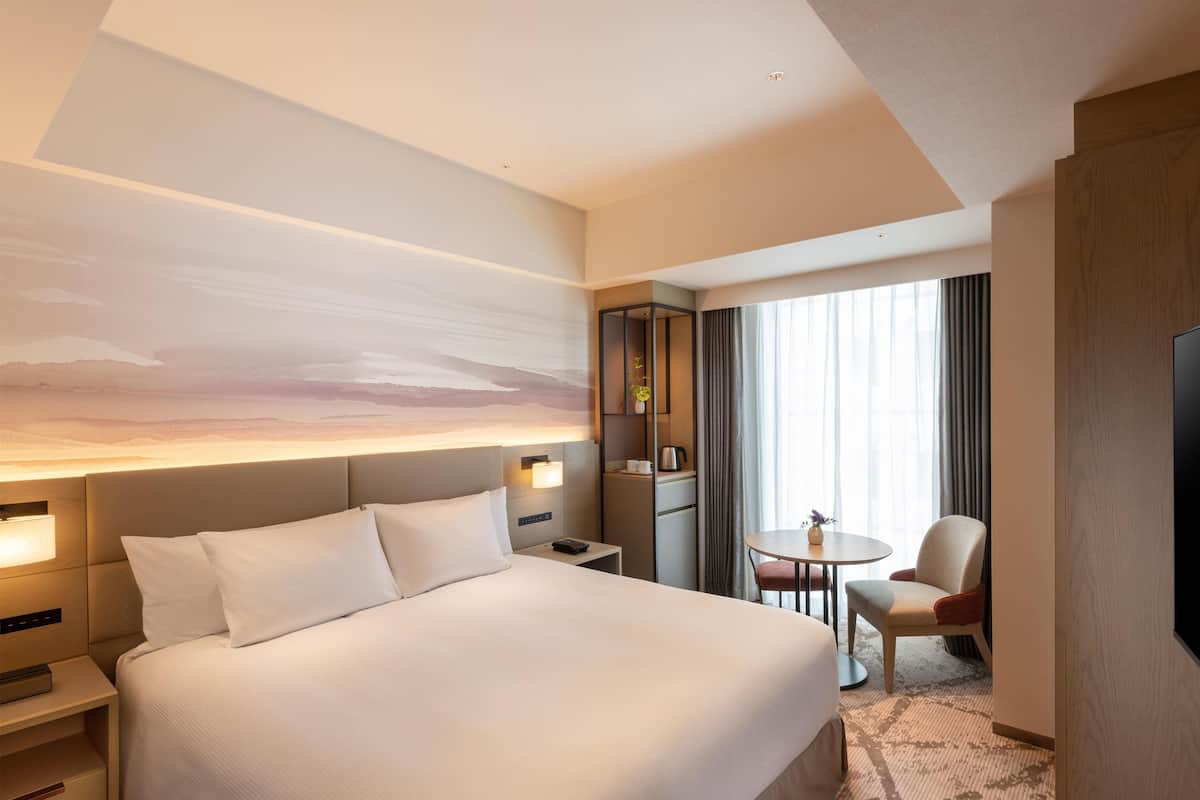 A hotel room with a large bed, bedside tables, a desk with a chair, a TV, and a window with curtains.