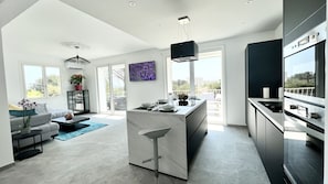 A modern kitchen with a central island, a dining area with a sofa and table, and a living room with a large window and artwork.