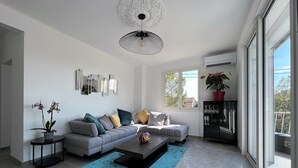 Ein modernes Wohnzimmer mit einem grauen Sofa, einem Couchtisch und einem blauen Teppich. Es befinden sich Blumentöpfe, ein wandmontierter Spiegel und eine Deckenleuchte dort.