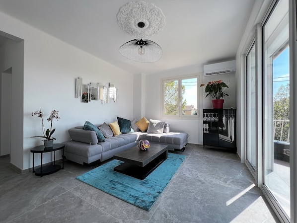 A modern living room with a grey sofa, a coffee table, and a blue rug. There are potted plants, a wall-mounted mirror, and a ceiling light fixture.