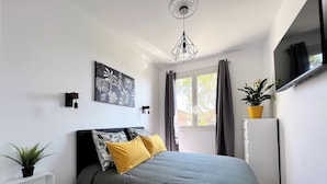 Ein Schlafzimmer mit Bett, einem an der Wand befestigten Fernseher, einer Zimmerpflanze und einem Fenster mit Vorhängen.