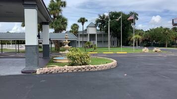 A parking area with a fountain, palm trees, and a building with a grey roof.
