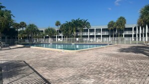 Uma área de piscina com piso de tijolos, cercada por um muro branco e palmeiras.