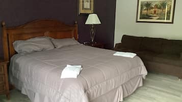 Una cama bien hecha con cabecera de madera, una mesita de noche con lámpara, un sofá marrón y dos cuadros enmarcados en la pared.