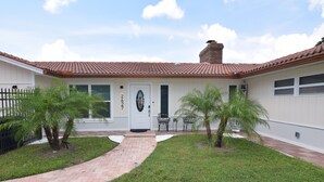 Una casa con techo de tejas, una chimenea de ladrillo y una puerta principal con placa de número. Hay dos palmeras y un camino pavimentado que conduce a la puerta.