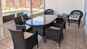 Un patio al aire libre con una mesa redonda y sillas de mimbre, un ventilador de techo y vistas a un jardín trasero con valla de madera y piscina.