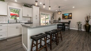 Een moderne keuken met witte kasten, een groot eiland met barstoelen, hanglampen en een eethoek met een tafel en stoelen.