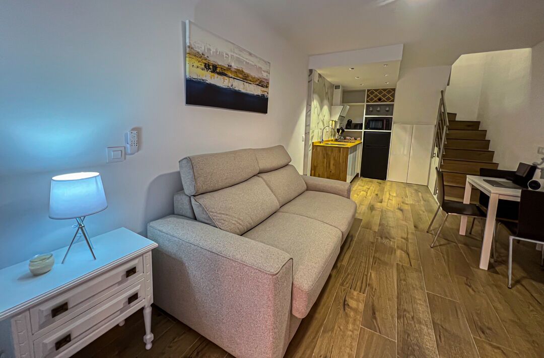 A modern living room with a sofa, a small table with a lamp, and a staircase leading to a kitchen area.