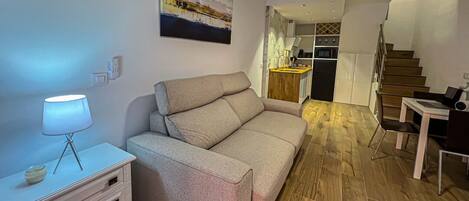 A modern living room with a sofa, a small table with a lamp, and a staircase leading to a kitchen area.