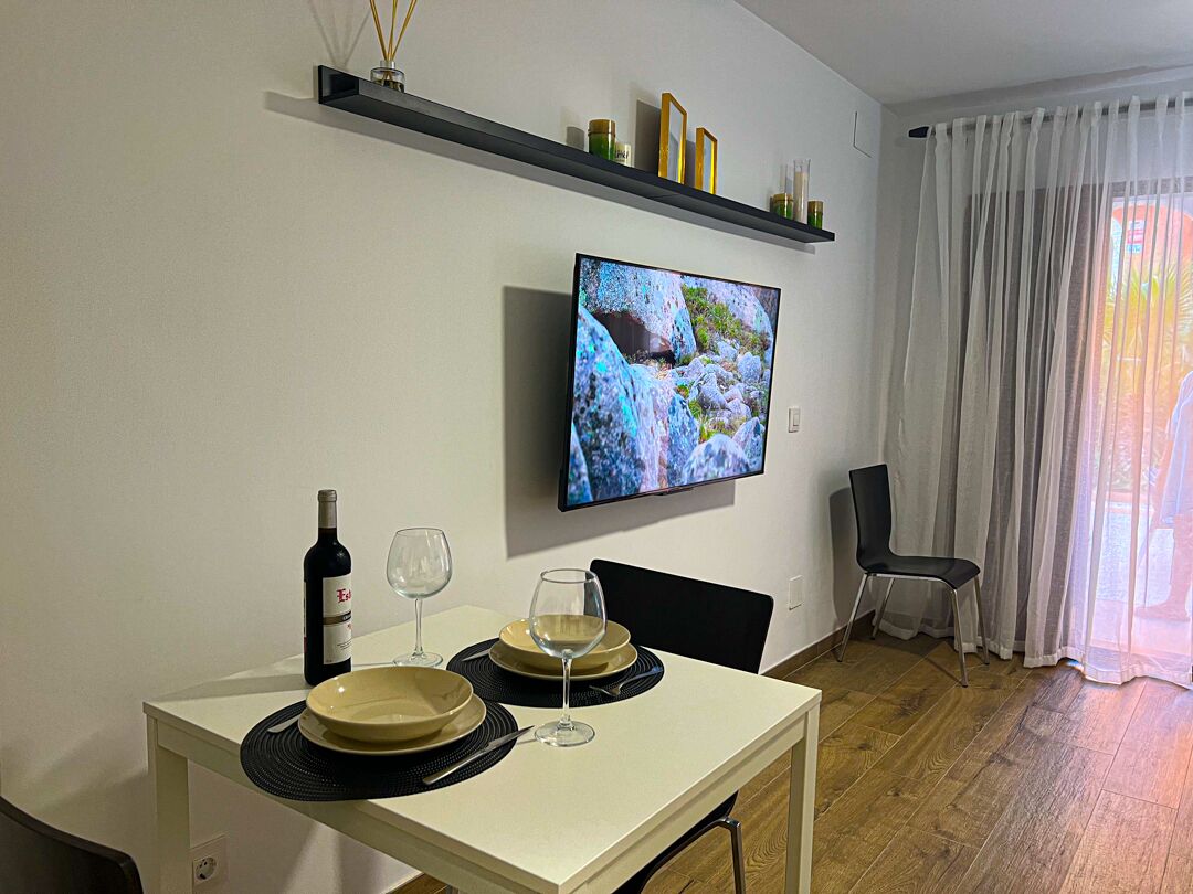 Un comedor con una mesa dispuesta para dos, un televisor montado en la pared y una ventana con cortinas de gasa.