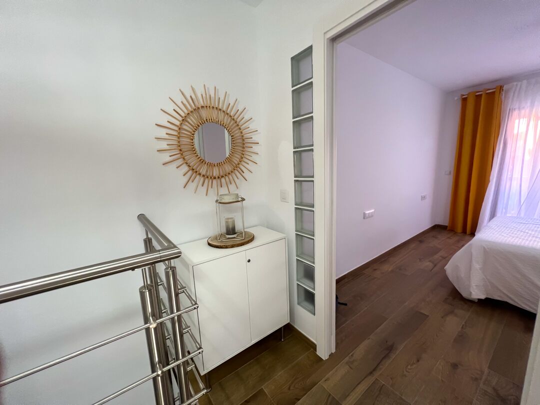 A modern bedroom with a wooden floor, a white bed, a glass cabinet, and a sunburst mirror.