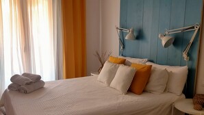 Ein ordentlich bezogenes Bett mit weißer Bettwäsche und einigen Kissen, ein Nachttisch mit einer Lampe sowie ein Fenster mit Vorhängen.