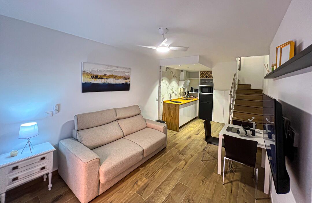 Un espacio habitable pequeño con sofá, una mesita, lámpara, ventilador de techo, área de cocina y una escalera.