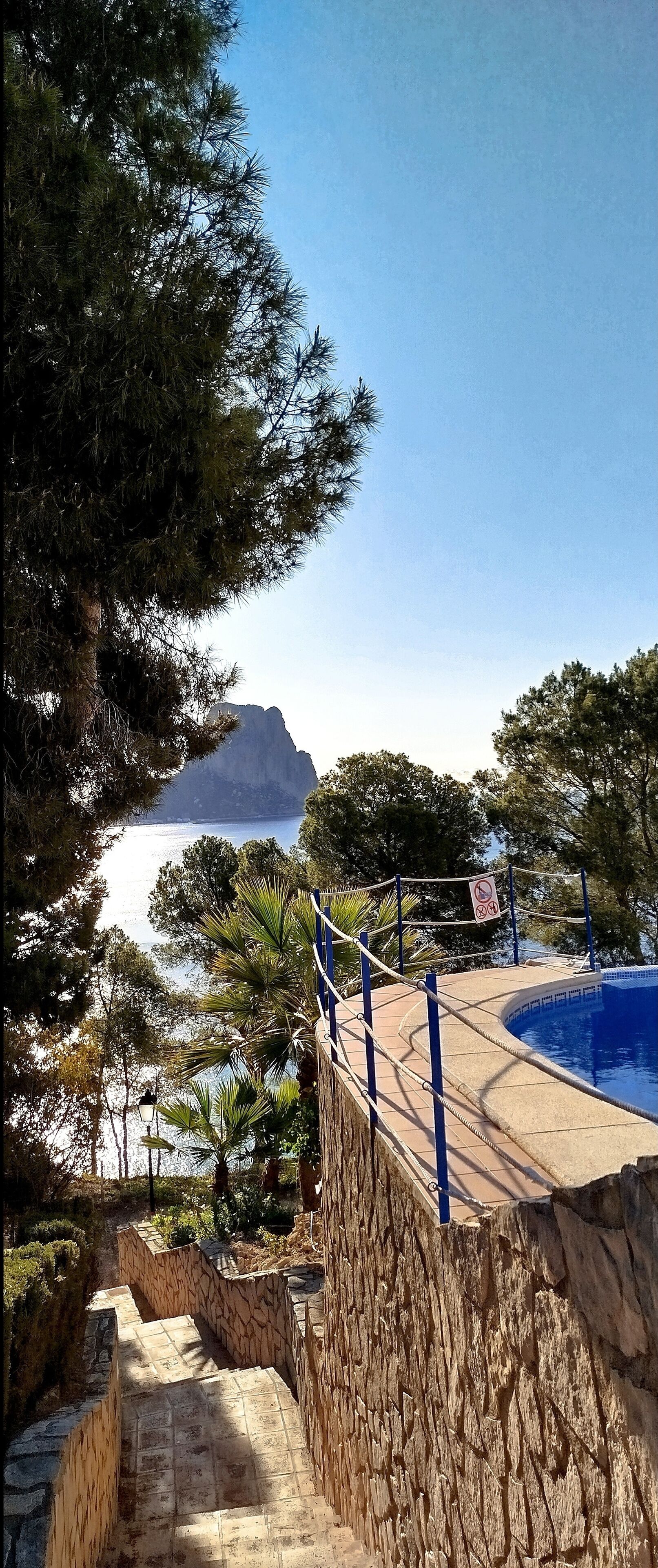 Un espace piscine agrémenté d’un mur en pierre, avec vue sur la mer et une montagne en arrière-plan.