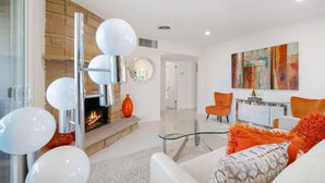A modern living room with a fireplace, a glass coffee table, orange chairs, and a contemporary art piece on the wall.