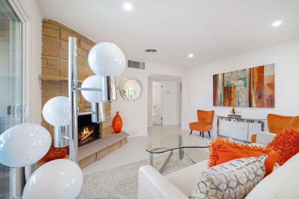 A modern living room with a fireplace, a glass coffee table, orange chairs, and a contemporary art piece on the wall.