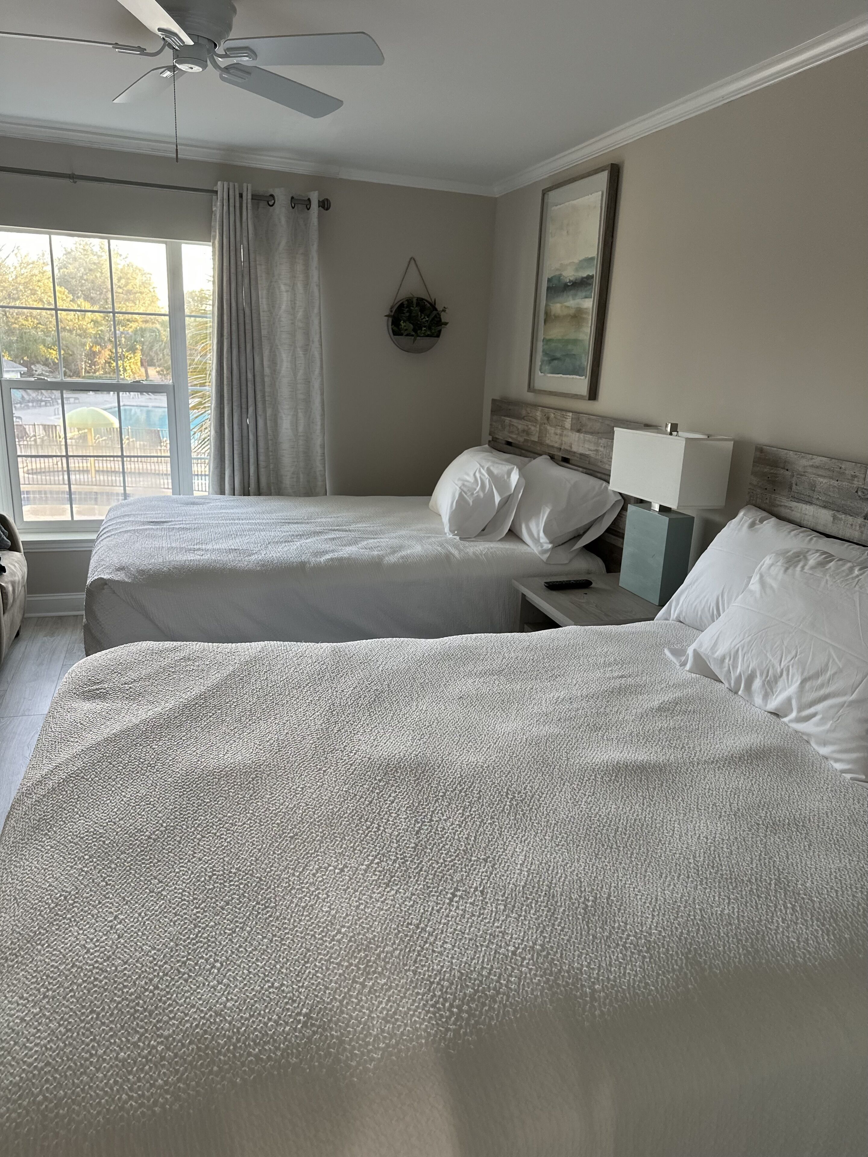 A hotel room with two beds, a ceiling fan, a window with curtains, a bedside table with a lamp, and a framed picture on the wall.