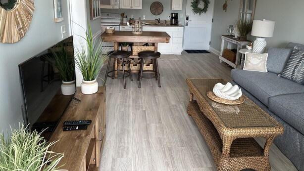 Una cocina moderna con armarios blancos, una isla central con taburetes y una zona de comedor con sofá gris y una mesita auxiliar de mimbre.